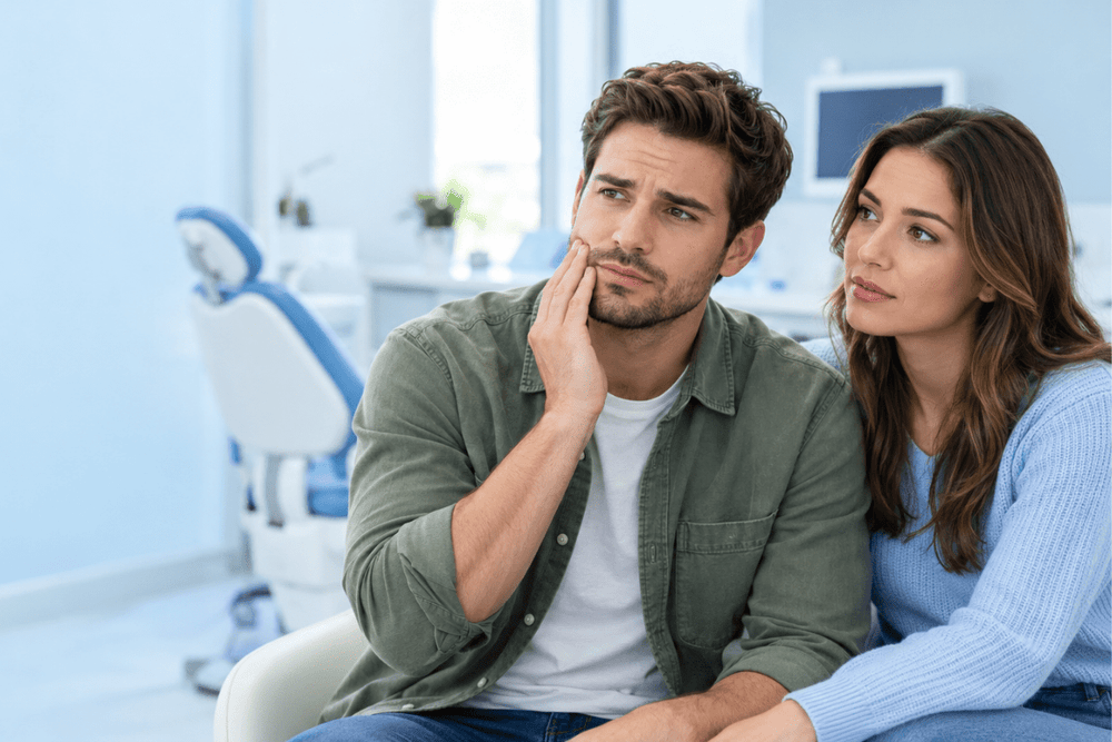 Man touching his jaw with a concerned expression while sitting beside a woman in a dental clinic.