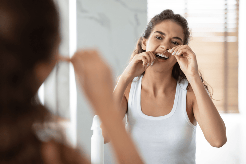 Woman flossing her teeth in front of a bathroom mirror before wearing a retainer