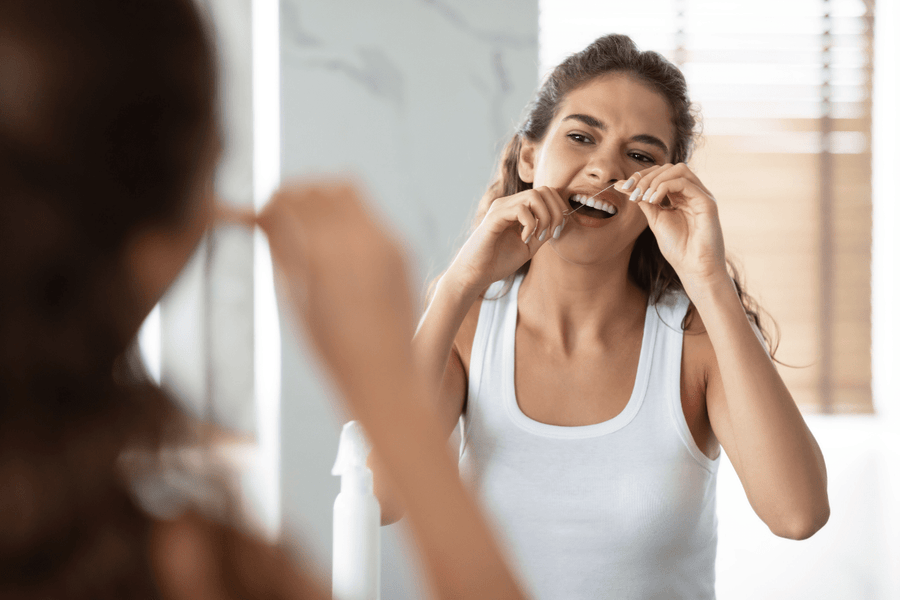 Woman flossing her teeth in front of a bathroom mirror before wearing a retainer