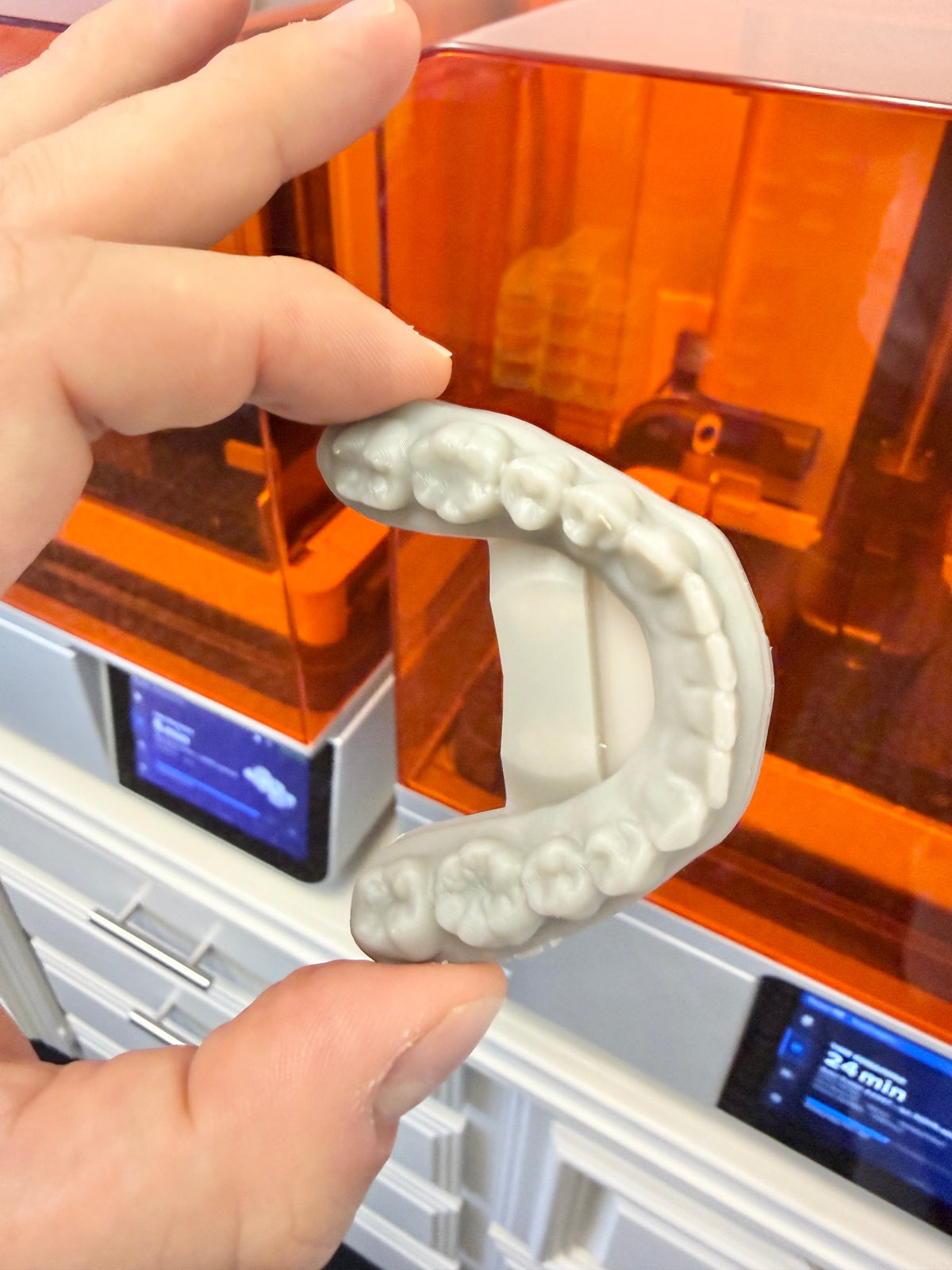A hand holds a light gray 3D-printed dental mold or impression in front of an orange and white 3D printer.