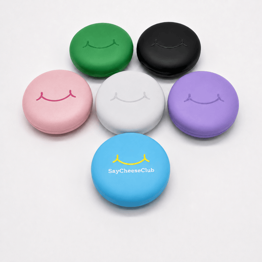 Six small round retainer silicone cases in different colors (green, black, pink, white, purple, and blue) arranged on a light gray background. Each case has a simple smiley face design on top, with the blue case featuring the text 'SayCheeseClub' and a yellow smiley graphic.