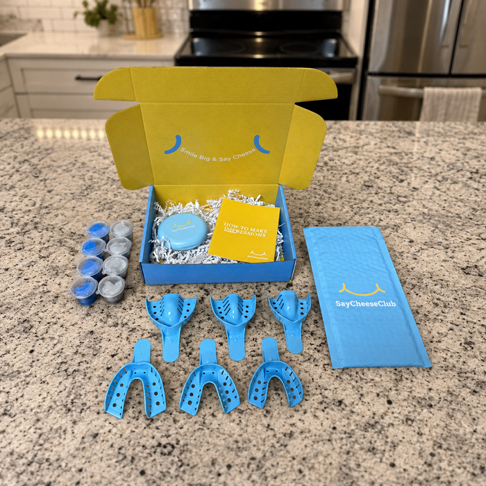 A dental mouth tray kit from SayCheeseClub arranged on a granite kitchen countertop, with the open branded box behind it and the blue trays, mouth pieces, pouch, and card in front.