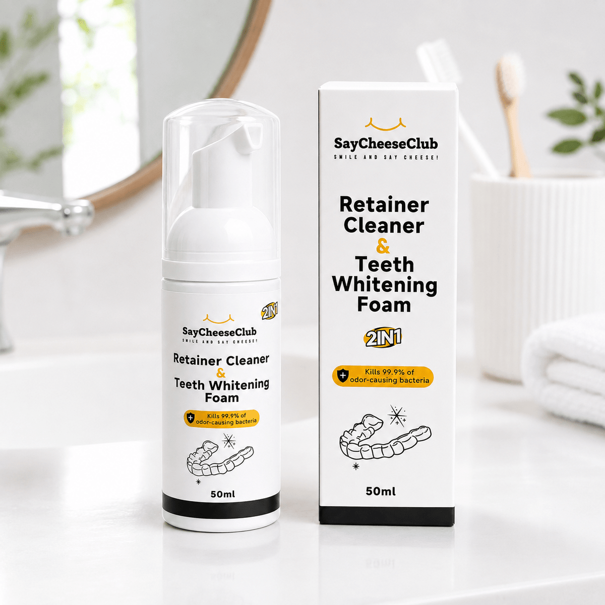 A 50ml bottle and matching box of SayCheeseClub Retainer Cleaner & Teeth Whitening Foam, set on a white bathroom counter with a toothbrush holder and sink in the background.