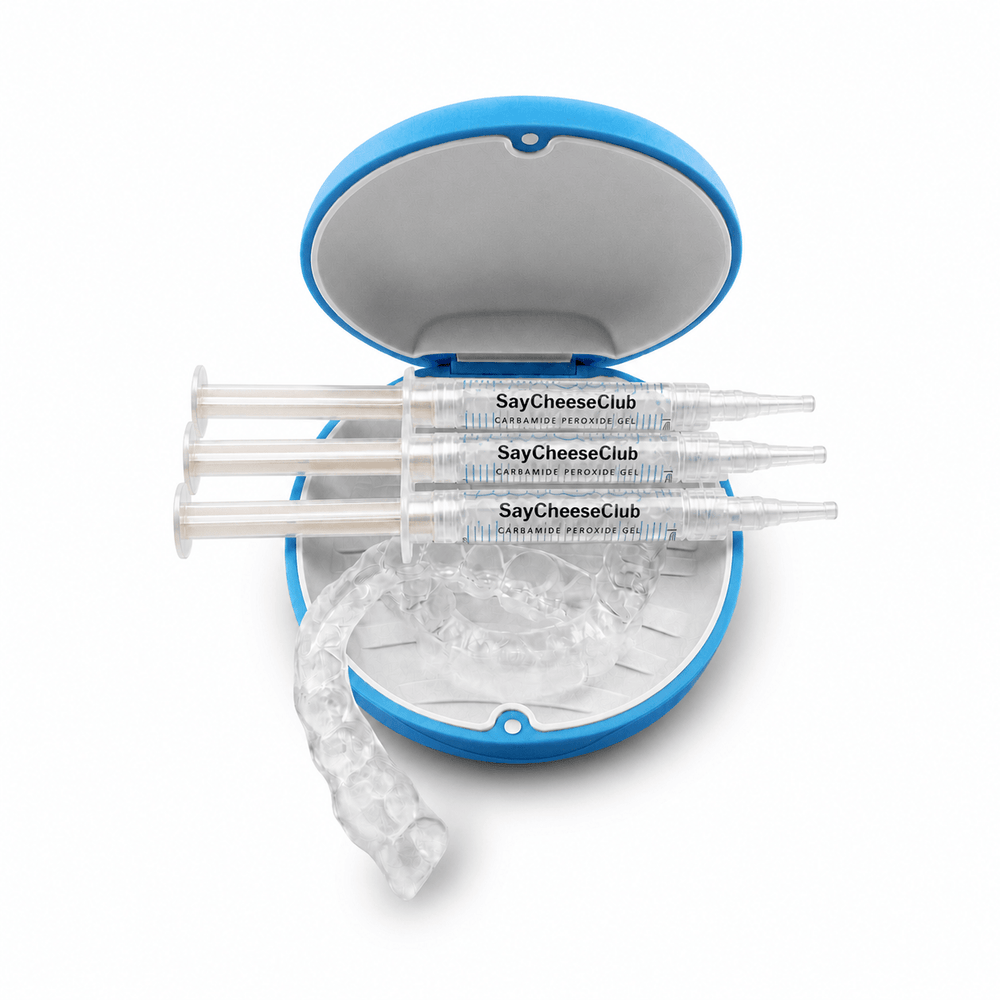 Three clear whitening tray syringe-style applicator tools labeled SayCheeseClub are arranged in front of an open blue-and-white round case, with a clear curved applicator piece resting in front on a white background.