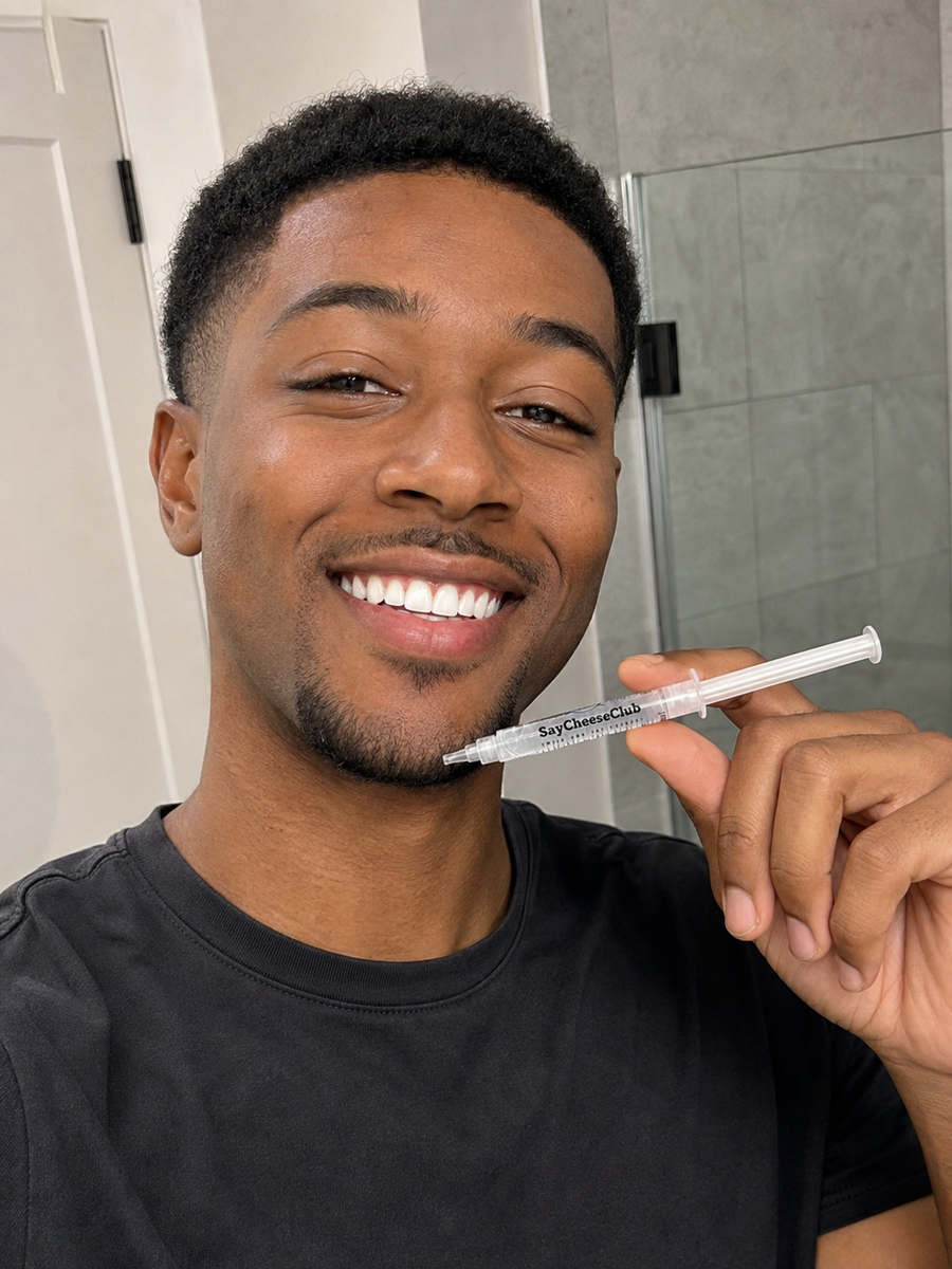 Smiling man in a black T-shirt holding a SayCheeseClub whitening gel syringe at an angle beside his chin in a bathroom.