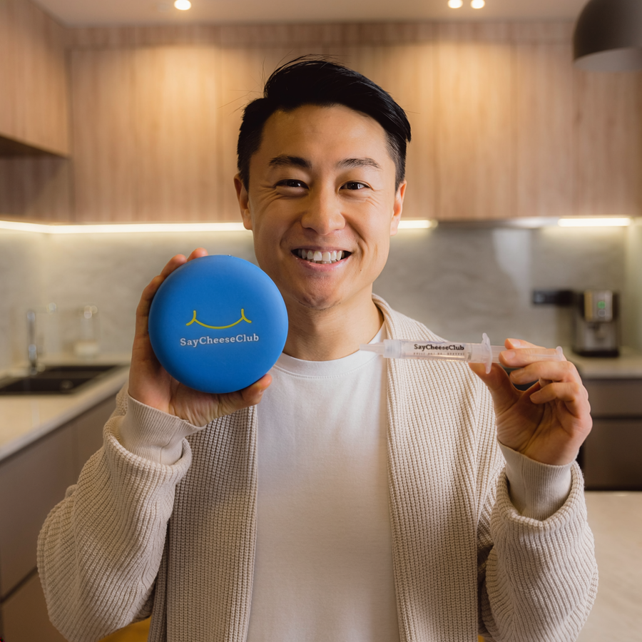 Smiling man holding a SayCheeseClub whitening gel syringe and blue case in a kitchen.