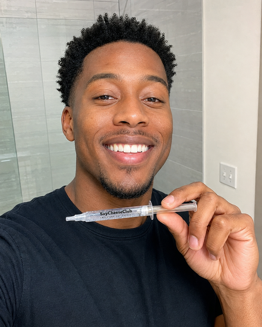 Smiling man in a black T-shirt holding a SayCheeseClub whitening gel syringe horizontally below his chin in a bathroom.