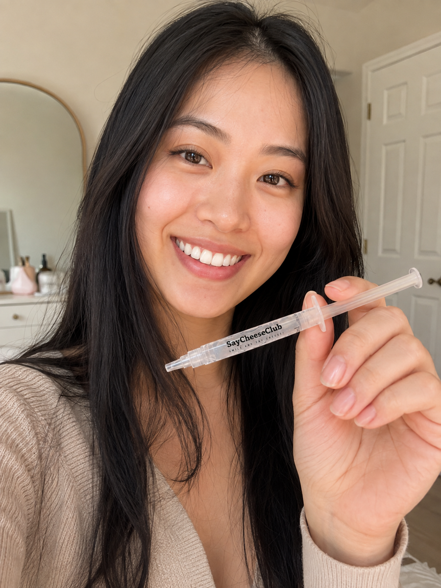 Smiling woman in a beige cardigan holding a SayCheeseClub whitening gel syringe, with a dresser and mirror behind her.