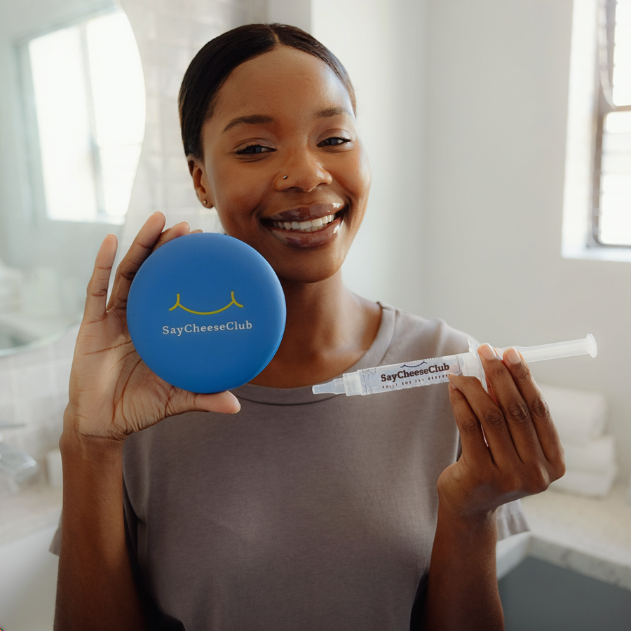 Smiling woman in a gray shirt holding a SayCheeseClub whitening gel syringe and blue case in a bathroom.