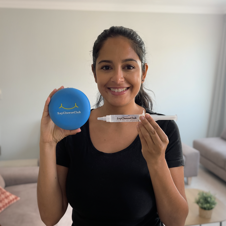 Smiling woman in a black shirt holding a SayCheeseClub whitening gel syringe and blue case in a living room.