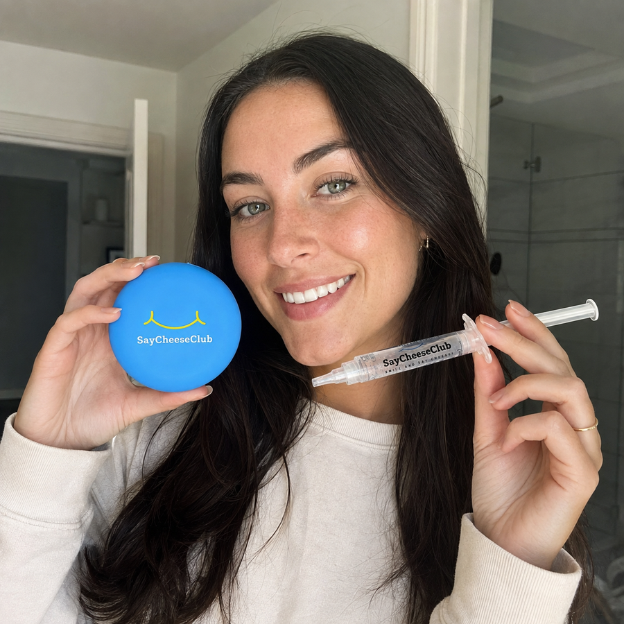 Smiling woman with long dark hair holding a SayCheeseClub whitening gel syringe and blue case in a bathroom.