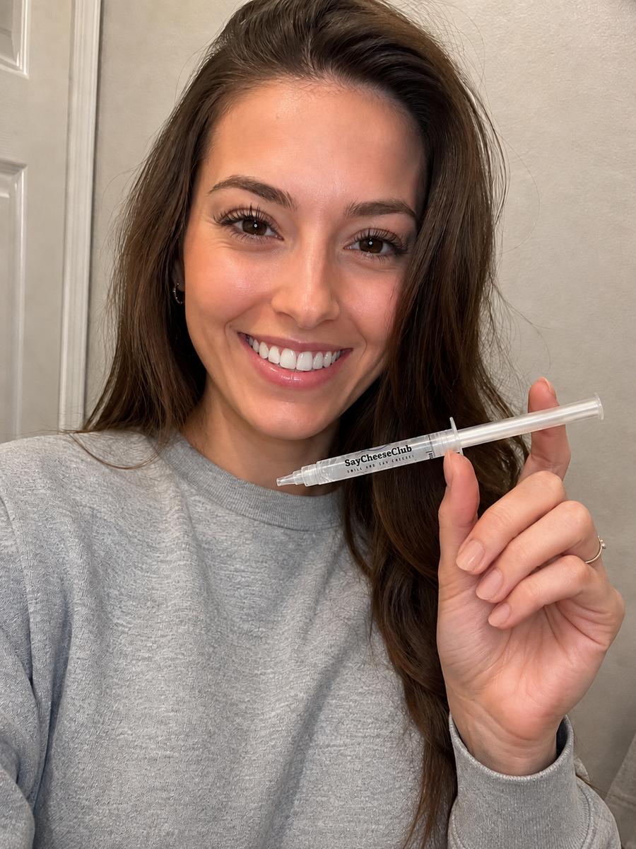 Smiling woman in a gray sweatshirt holding a SayCheeseClub whitening gel syringe beside her face against a light wall.
